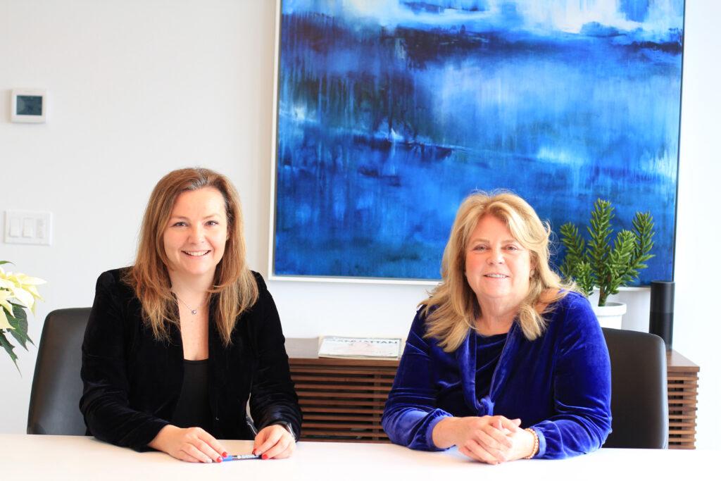 two women in formal clothing sitting at a table and smiling at the camera with their hands folded