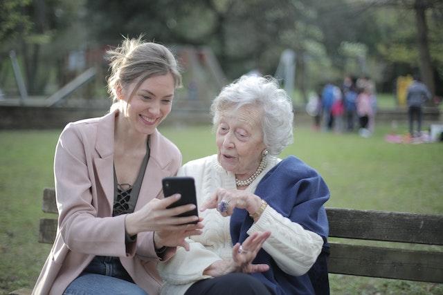 a young woman showing her phone to an elderly woman who is sitting next to her on the same bench