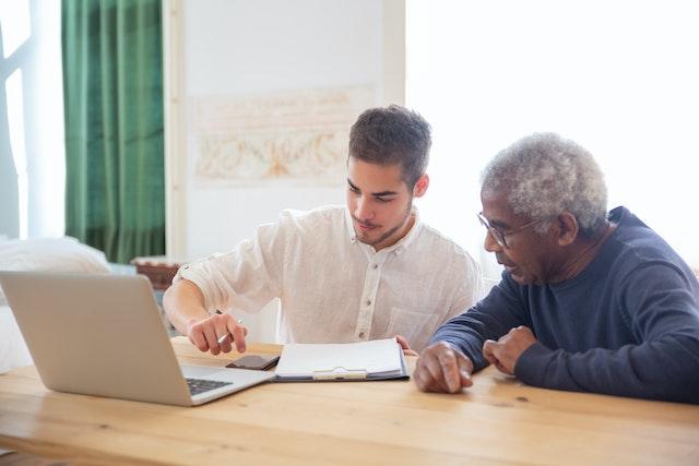 a man sitting next to an elderly man at a desk and using a calculator in front of an open laptop