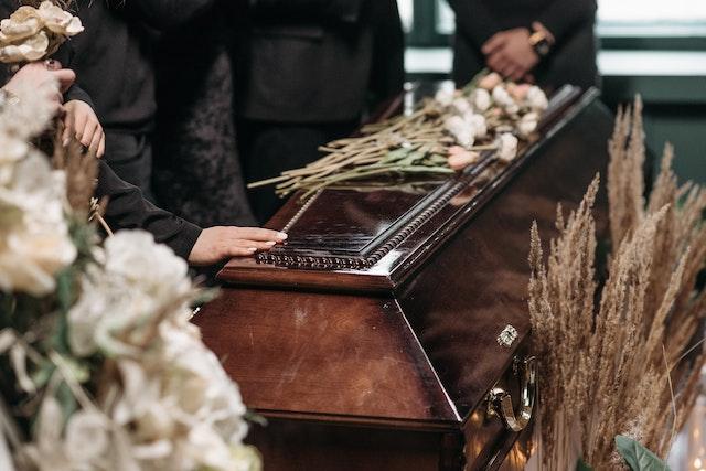a person placing their hand on a closed, wooden casket with flowers on it