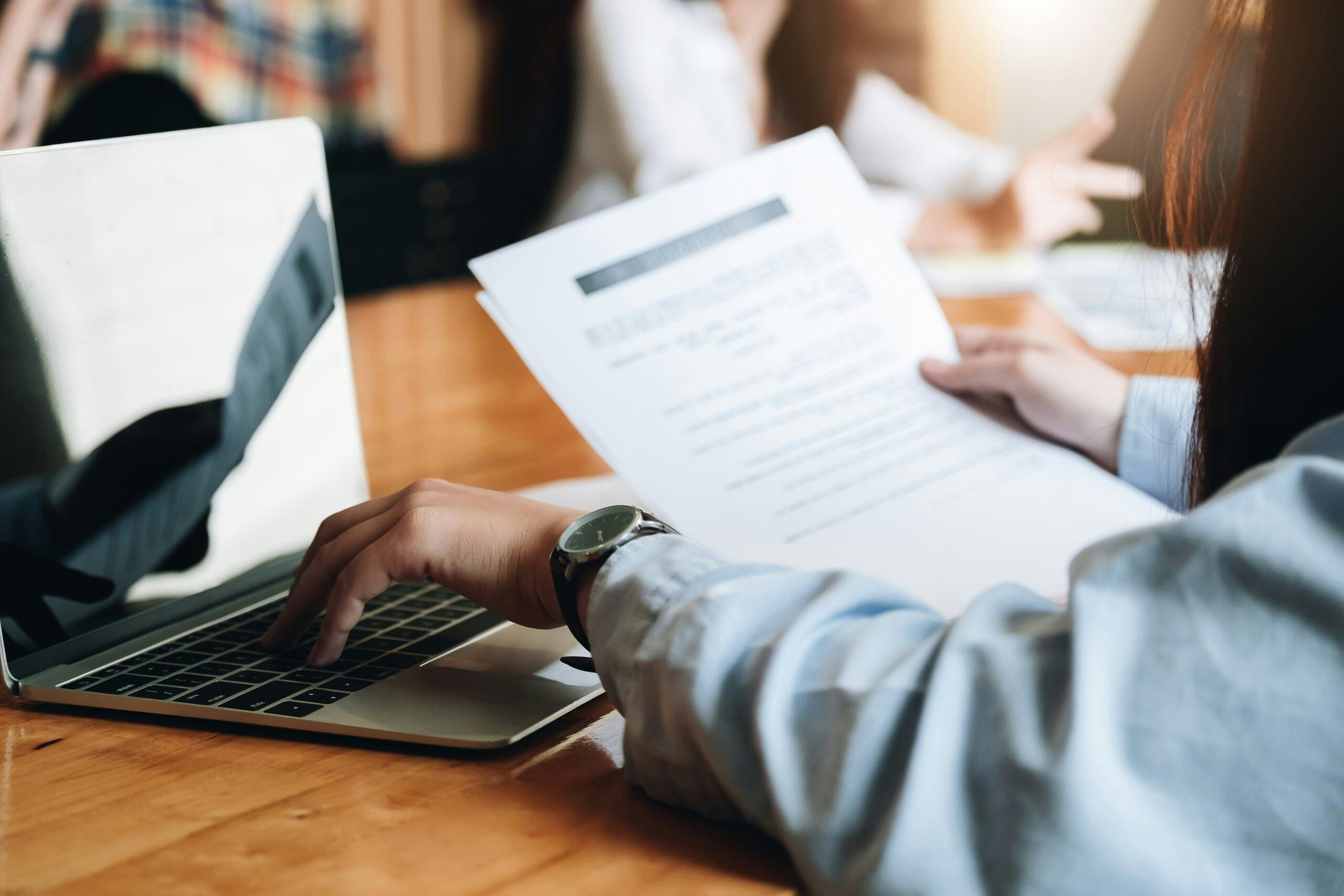 a person holding a paper document in one hand and typing on a laptop with the other